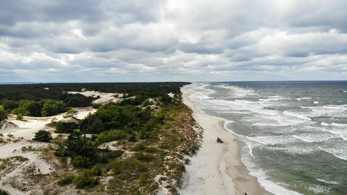 Ein Drohnenblick auf die Halbinsel Hel zeigt den langen Sandstrand, der sich entlang der Küste der polnischen Ostsee erstreckt. Das klare, dunkelblaue Wasser trifft auf die sanften Dünen und den angrenzenden Kiefernwald, der die Landschaft prägt. Ein harmonisches Zusammenspiel von Natur und Küstenlandschaft.