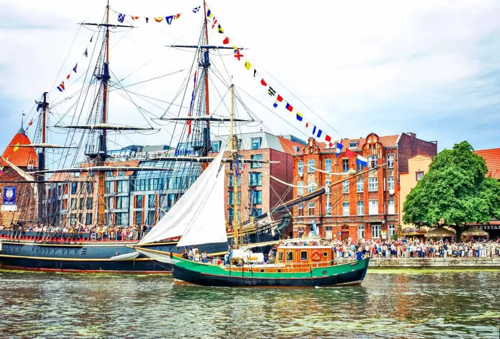 Kleines Segelboot im Vordergrund vor einem großen historischen Segelschiff auf der Motława in Danzig, mit vielen Besuchern an den Uferpromenaden.