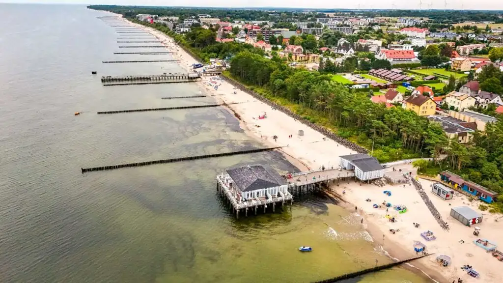Luftaufnahme von Ustronie Morskie mit zwei Seebrücken, grün-blauer Ostsee und dem dahinter liegenden Ort an der polnischen Ostseeküste.